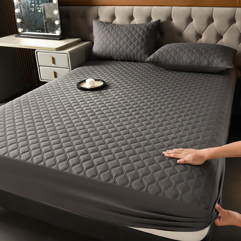 A dark gray quilted waterproof mattress protector is shown on a bed with matching pillows and a small tray.
