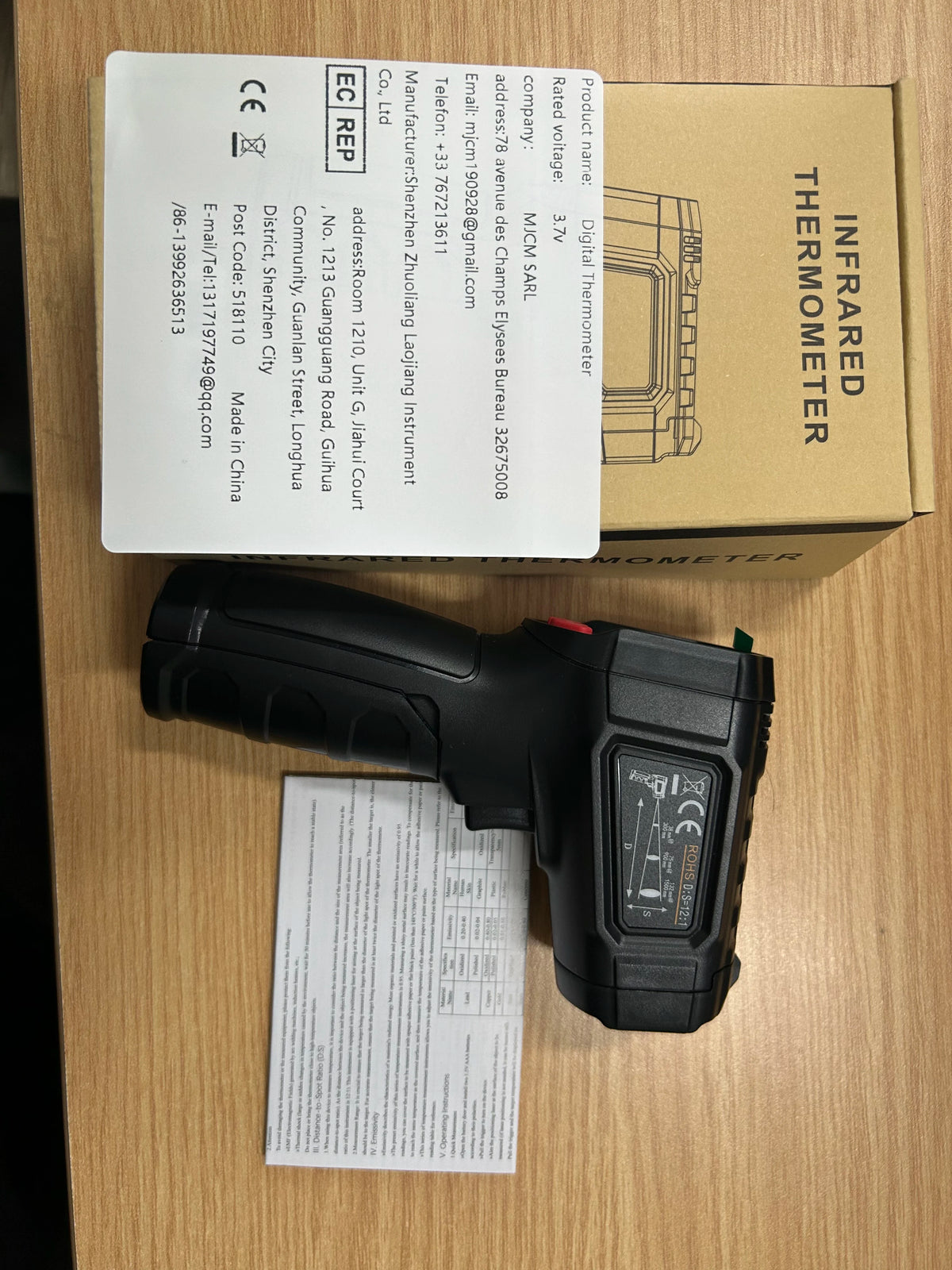 A black digital infrared thermometer is shown next to its packaging and a product information sheet.