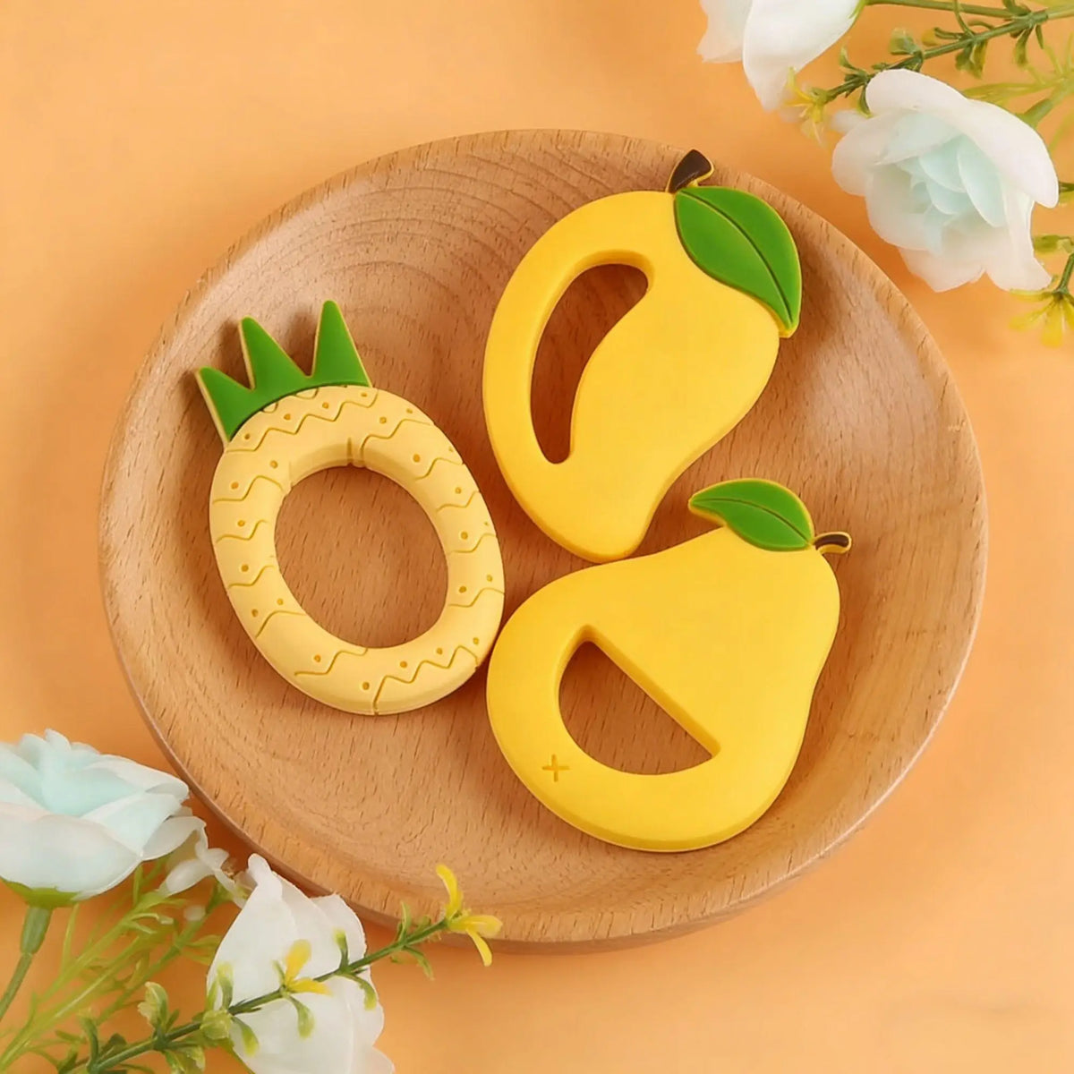 A yellow silicone fruit-shaped teether, resembling a lemon or pear, rests on a wooden plate with white flowers.