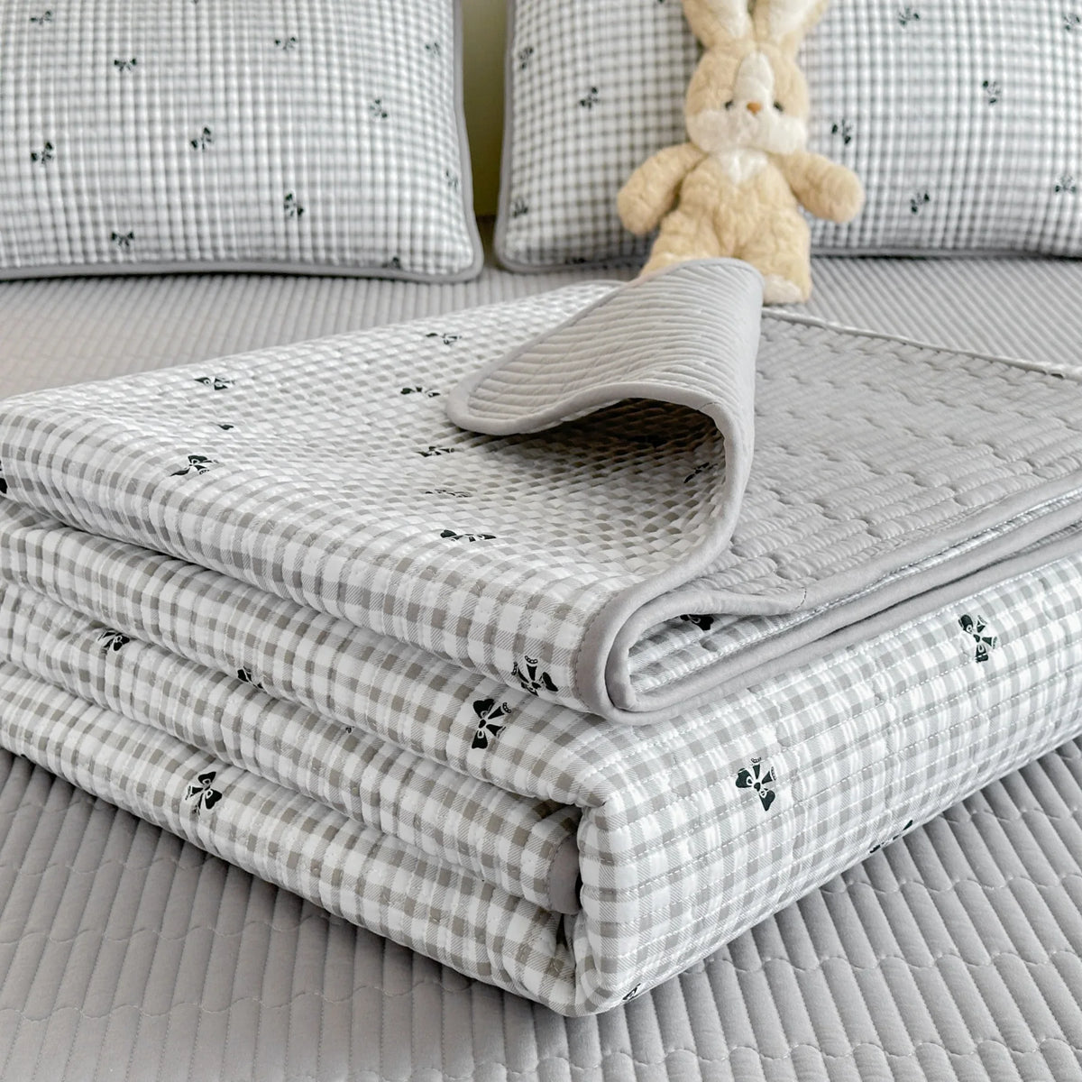 A stack of light gray floral quilted linens and a small cream stuffed rabbit toy on a bed.