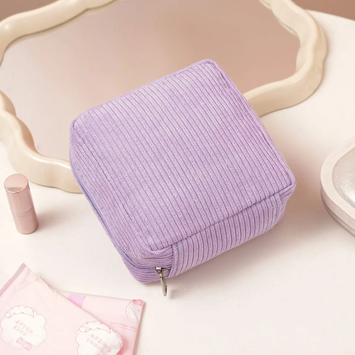A lavender ribbed storage pouch with a zipper is displayed on a white surface next to a mirror frame and pink sanitary napkin