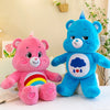 Un peluche de osito de cuidado arcoíris rosa sentado junto a un peluche de oso azul.