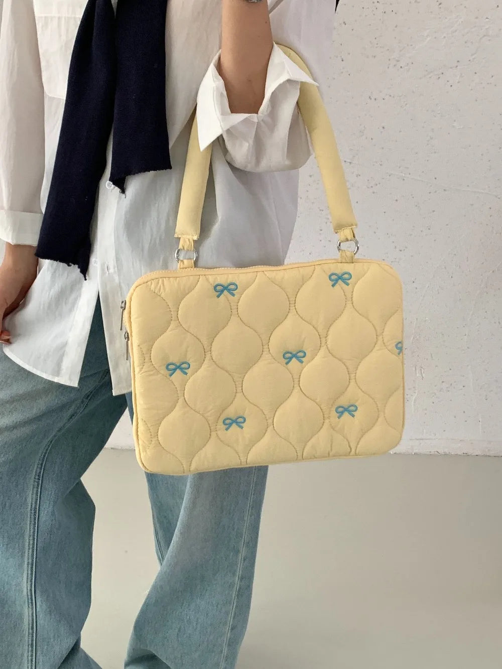 A light yellow quilted laptop shoulder bag with blue bows is carried by a person wearing a white shirt and jeans.