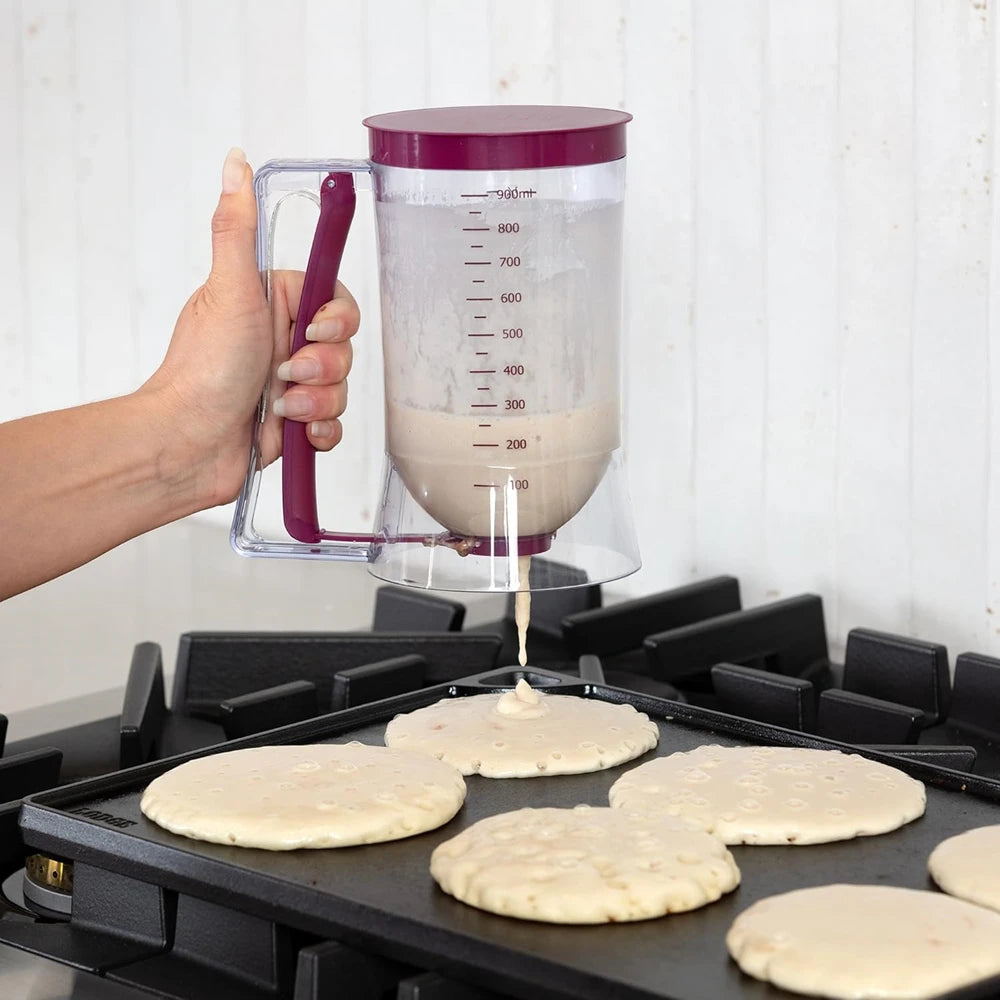 A hand pours pancake batter into a clear dispenser with a measuring guide onto a baking sheet.