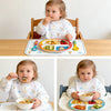 Two babies eating food from plates and bowls on a wooden table.