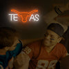 A young man wearing an orange jersey is smiling while a neon sign shaped like a cow head spelling TEAS is visible in the back