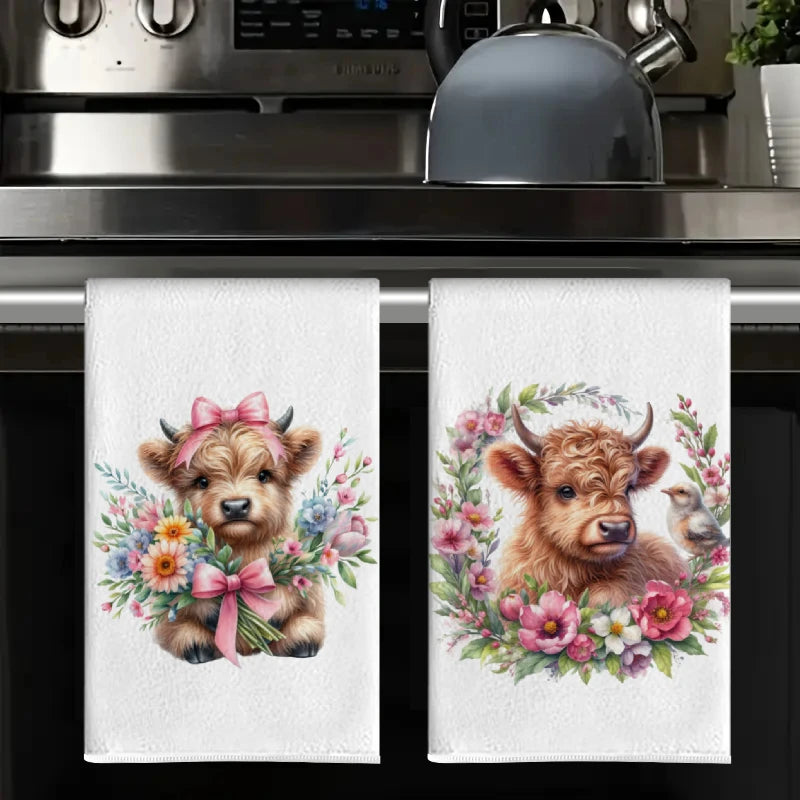 A set of two white kitchen towels featuring watercolor illustrations of cute Highland cows with floral wreaths.