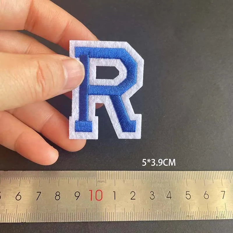 A hand holds a blue embroidered letter R patch against a ruler showing its size.