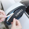 A hand is removing a carbon fiber rain guard from a white car's rearview mirror.