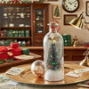 A glass bottle shaped like a Christmas tree with glitter and ornaments sits on a gold plate in a festive holiday setting.
