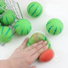 Eine Hand drückt ein wassermelonenförmiges Stressabbau-Spielzeug, umgeben von ähnlichen Spielzeugen und einem Drahtkorb.