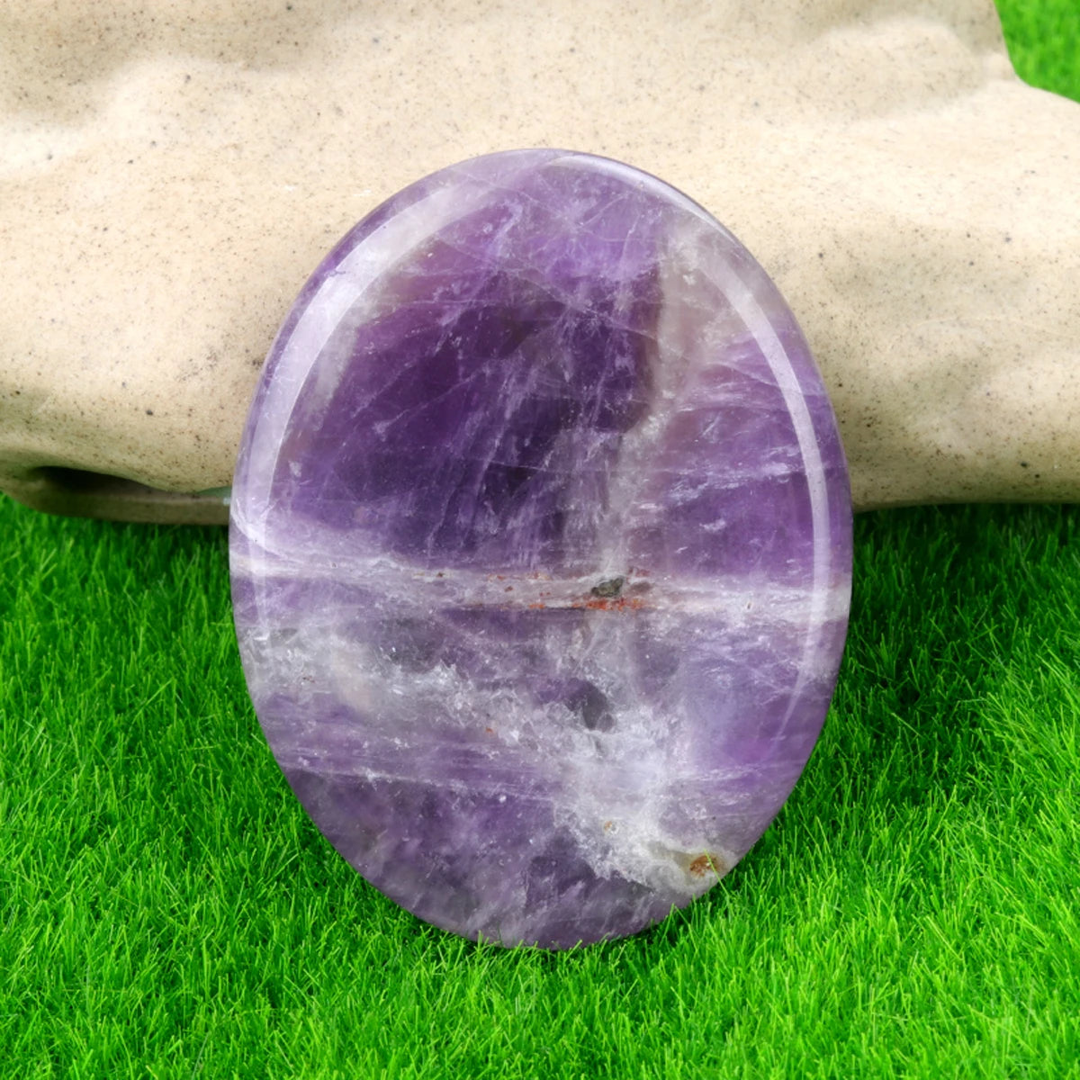 A smooth, oval purple amethyst crystal stone resting on green grass, held by a hand.