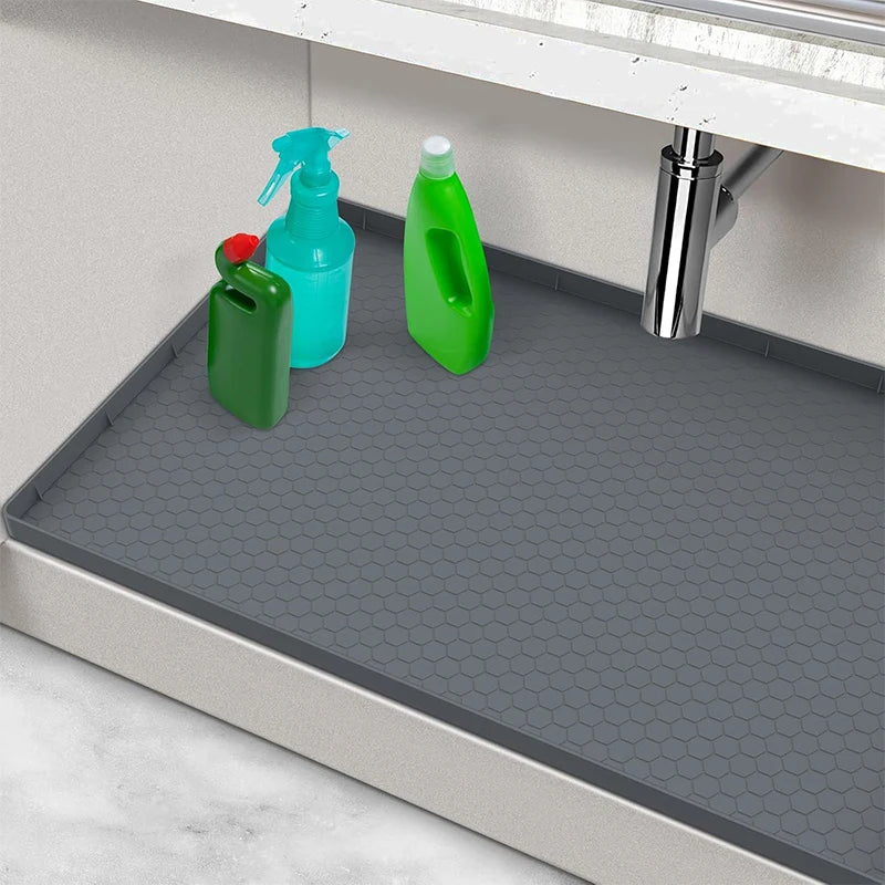 A flexible silicone under-sink mat with a raised edge is shown in a kitchen context with various cleaning bottles.