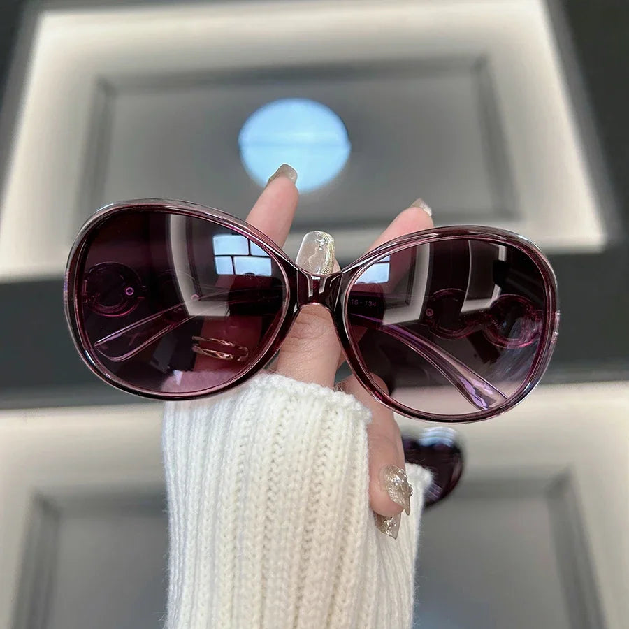 A hand holds a pair of square vintage sunglasses with dark purple lenses and gold accents against a bright background.