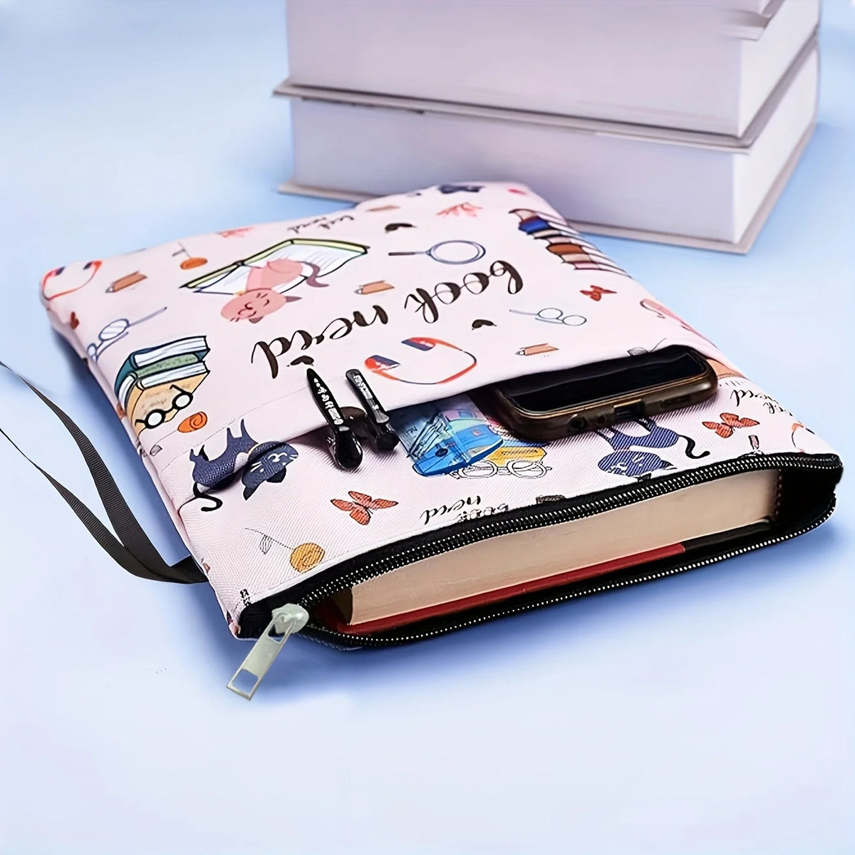 A pink fabric book sleeve with a zipper features a cat and books design, resting on a light blue surface.