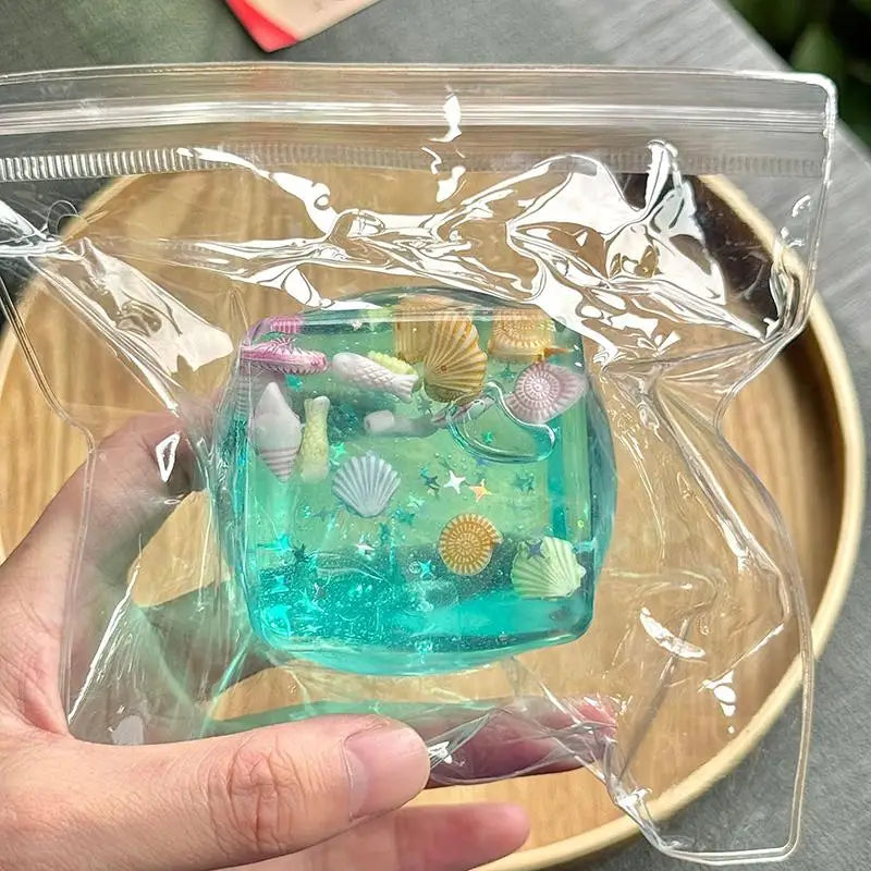 A hand holds a clear plastic bag containing a turquoise ocean-themed squeeze toy with seashells and sea life.