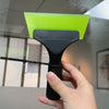 A hand holds a black ice scraper with a bright lime green plastic squeegee blade against a glass surface.