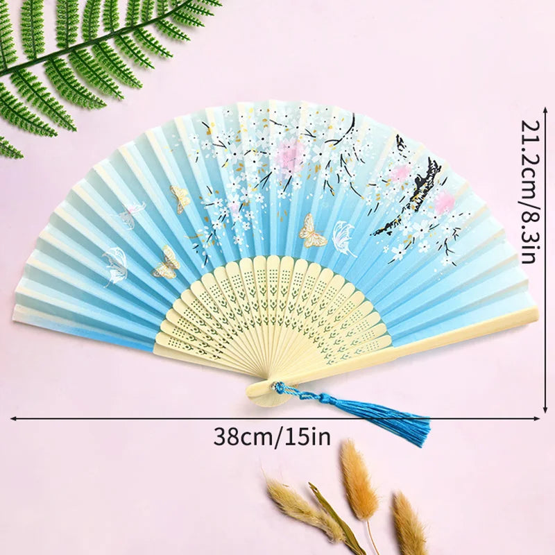 A light blue folding hand fan decorated with floral and butterfly patterns, shown against a pink background.