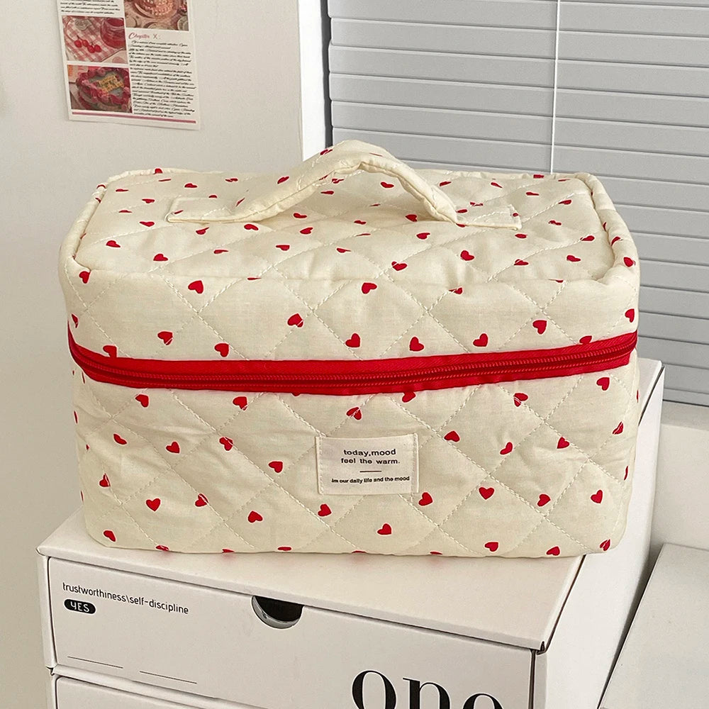 A cream-colored quilted makeup bag with a red piping detail and a heart pattern is resting on a white box.