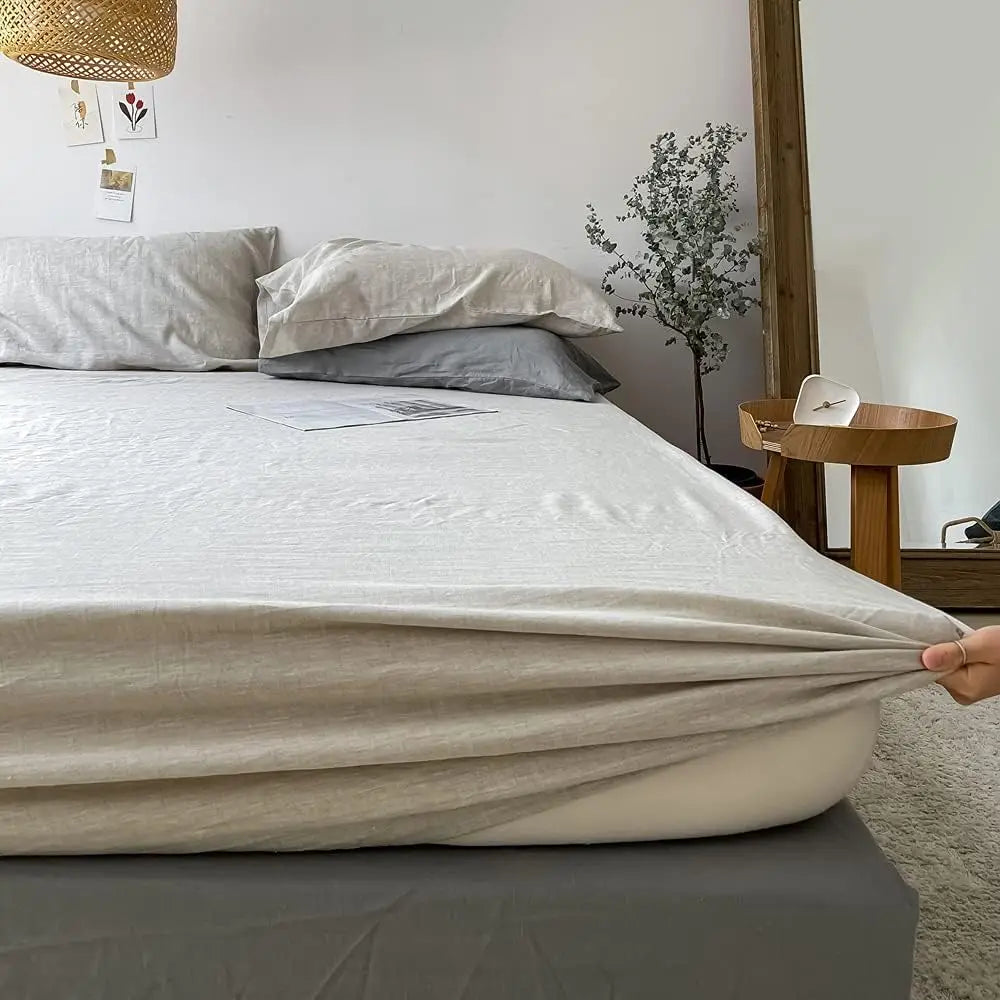 A white linen fitted sheet is laid out on a bed with a gray duvet and pillows.