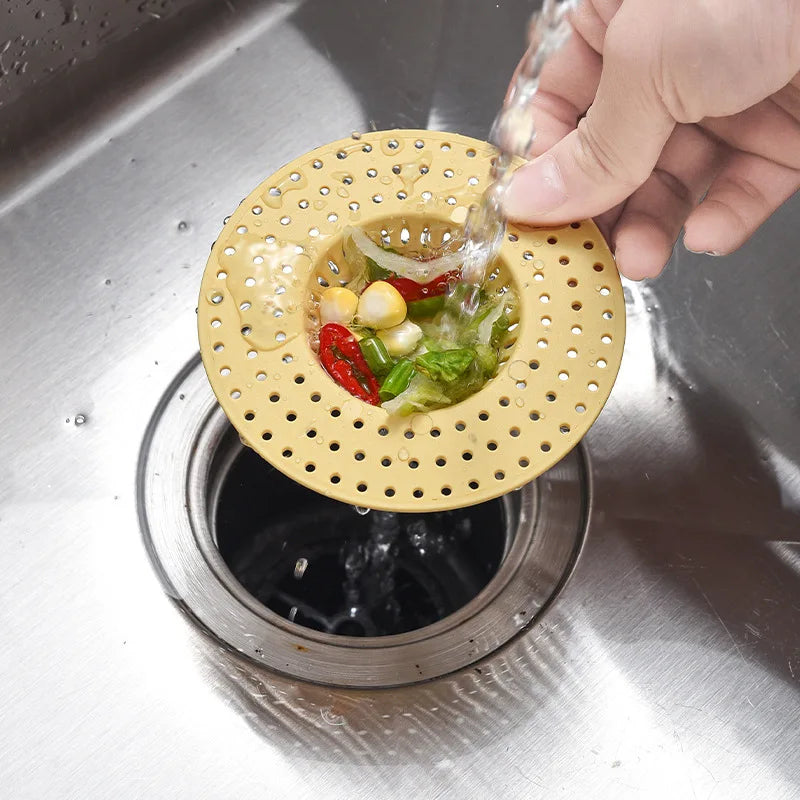 A hand is pouring water through a yellow silicone sink drain filter strainer into a metal sink.