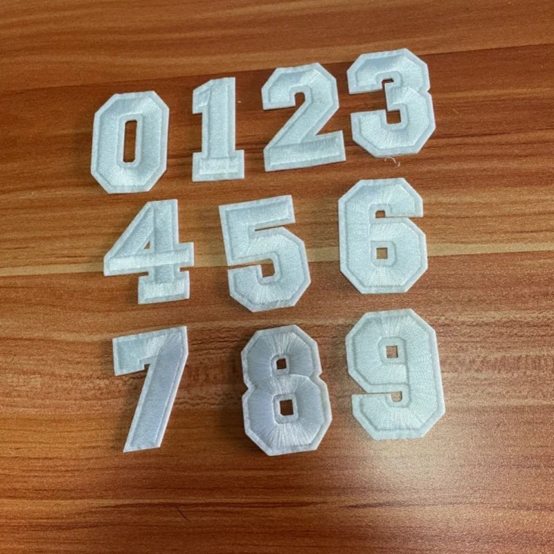 A set of white iron-on 3D number patches from 0 to 9 laid out on a wooden surface.