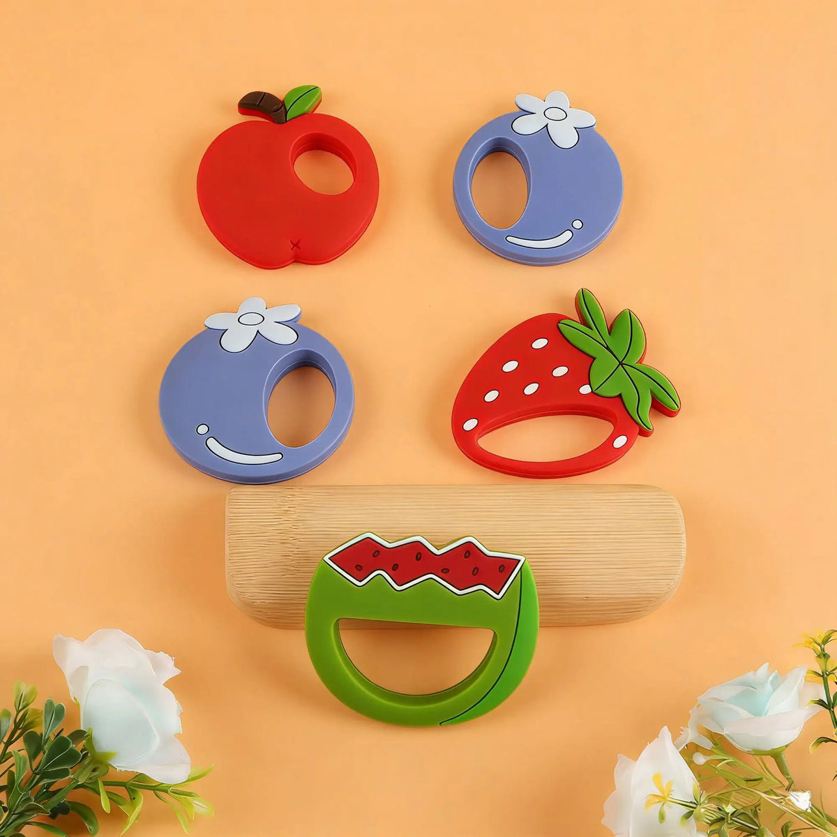 A set of silicone fruit-shaped baby teethers, including an apple and a strawberry, are displayed on a light orange background