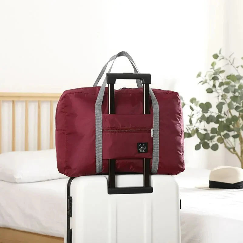 A maroon foldable travel duffel bag with gray straps resting on a white rolling suitcase.