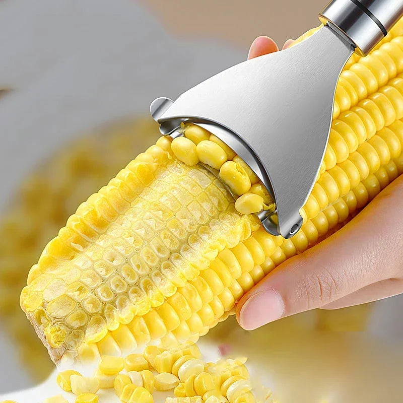 A hand is using a stainless steel corn peeler to remove kernels from a corn cob.