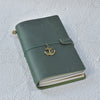 A dark olive green leather refillable journal with a brass anchor charm attached to the cover.