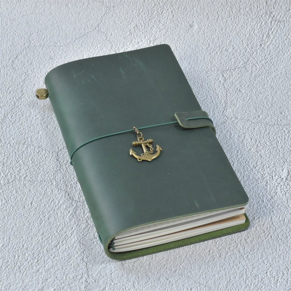 A dark olive green leather refillable journal with a brass anchor charm attached to the cover.