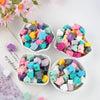 A collection of colorful silicone crown star heart beads in various shades are displayed in small white bowls on a white back