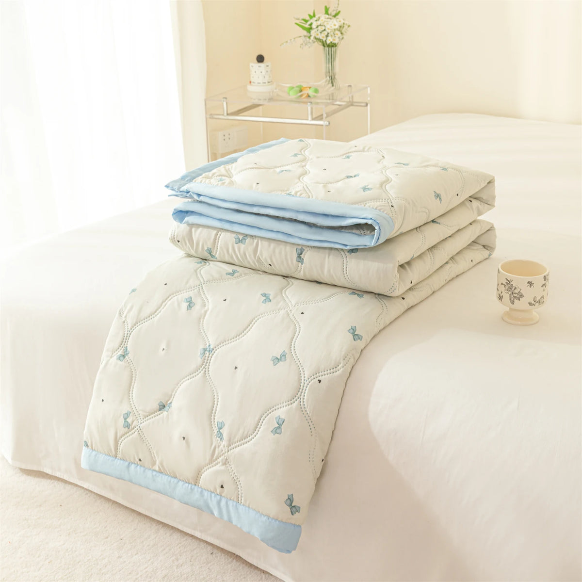 A light blue quilted summer quilt with small butterfly embroidery is folded on a white bed.