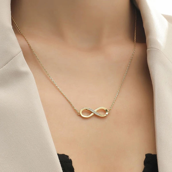 Un collier pendentif à l'infini de couleur or est présenté sur un fond neutre, porté autour du cou sous un vêtement clair