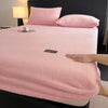 A pink plush mattress cover is displayed on a bed, next to a lamp and pillows.