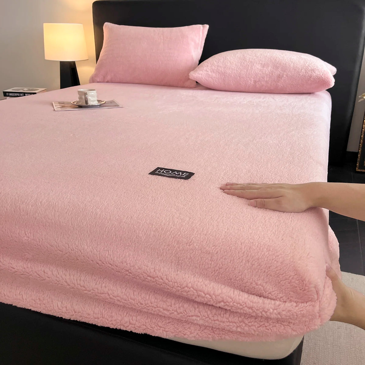 A pink plush mattress cover is displayed on a bed, next to a lamp and pillows.