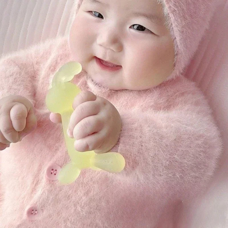 A baby is holding a yellow silicone teether while wearing a pink fuzzy hooded garment.