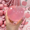 Eine Hand hält ein durchsichtiges, apfelförmiges Fidget-Spielzeug mit Blatt darauf vor einem Hintergrund aus rosa Blumen und 
