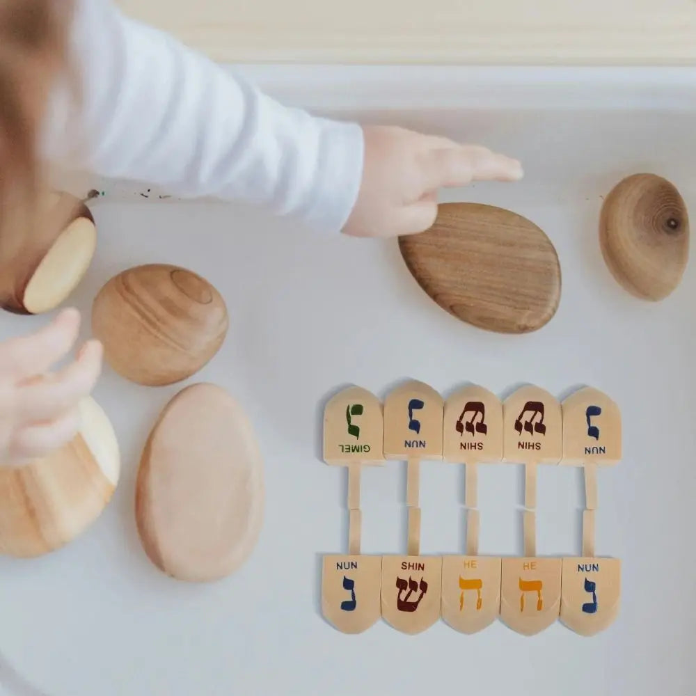 A hand places wooden spinning tops labeled with Hebrew letters onto a white surface.
