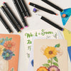 A collection of black and colorful dual-tip alcohol markers and art paper on a white surface.