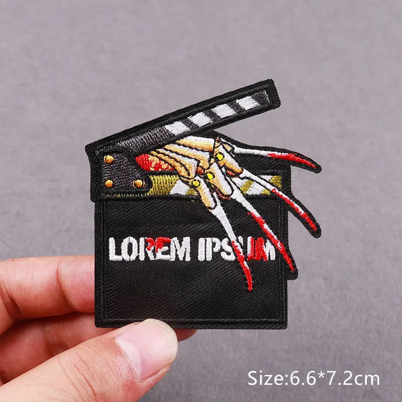 Black embroidered patch featuring a stylized skull with sharp teeth and dripping blood, displayed on a hand.