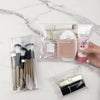 A clear cosmetic bag contains makeup brushes and a gold-colored cosmetic product on a white marble surface.