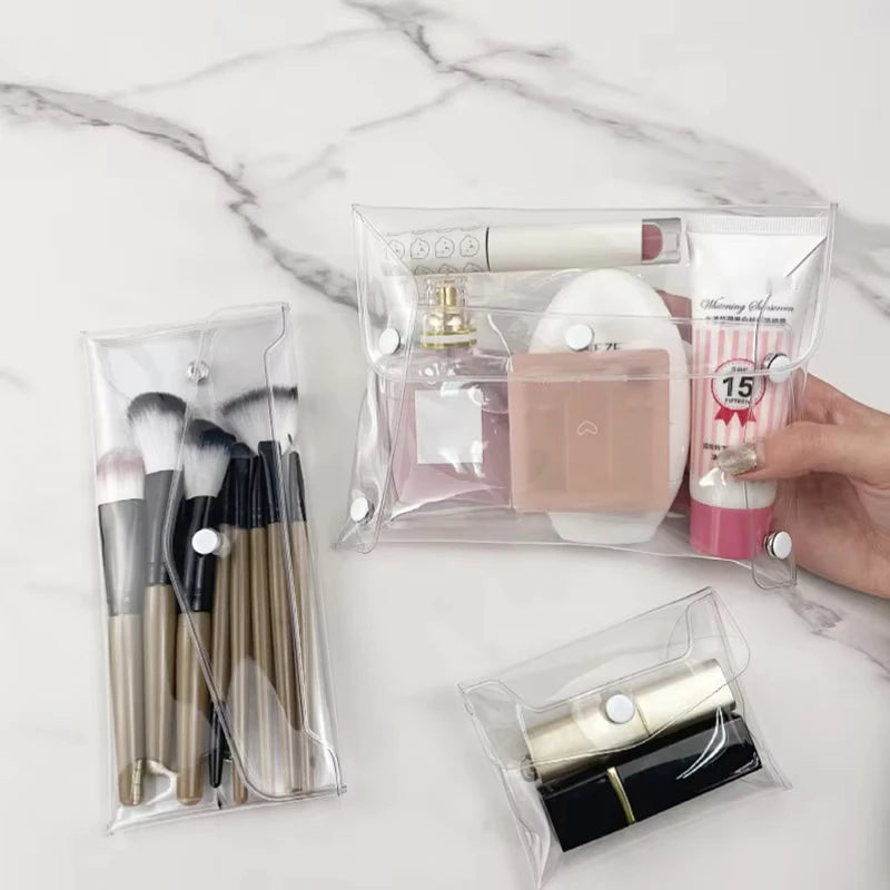 A clear cosmetic bag contains makeup brushes and a gold-colored cosmetic product on a white marble surface.