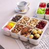 A clear portable snack container filled with various nuts, dried fruits, and grapes, set next to a teacup.