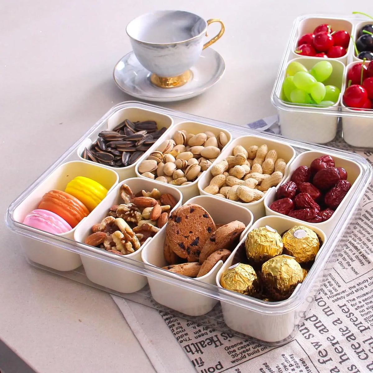 A clear portable snack container filled with various nuts, dried fruits, and grapes, set next to a teacup.
