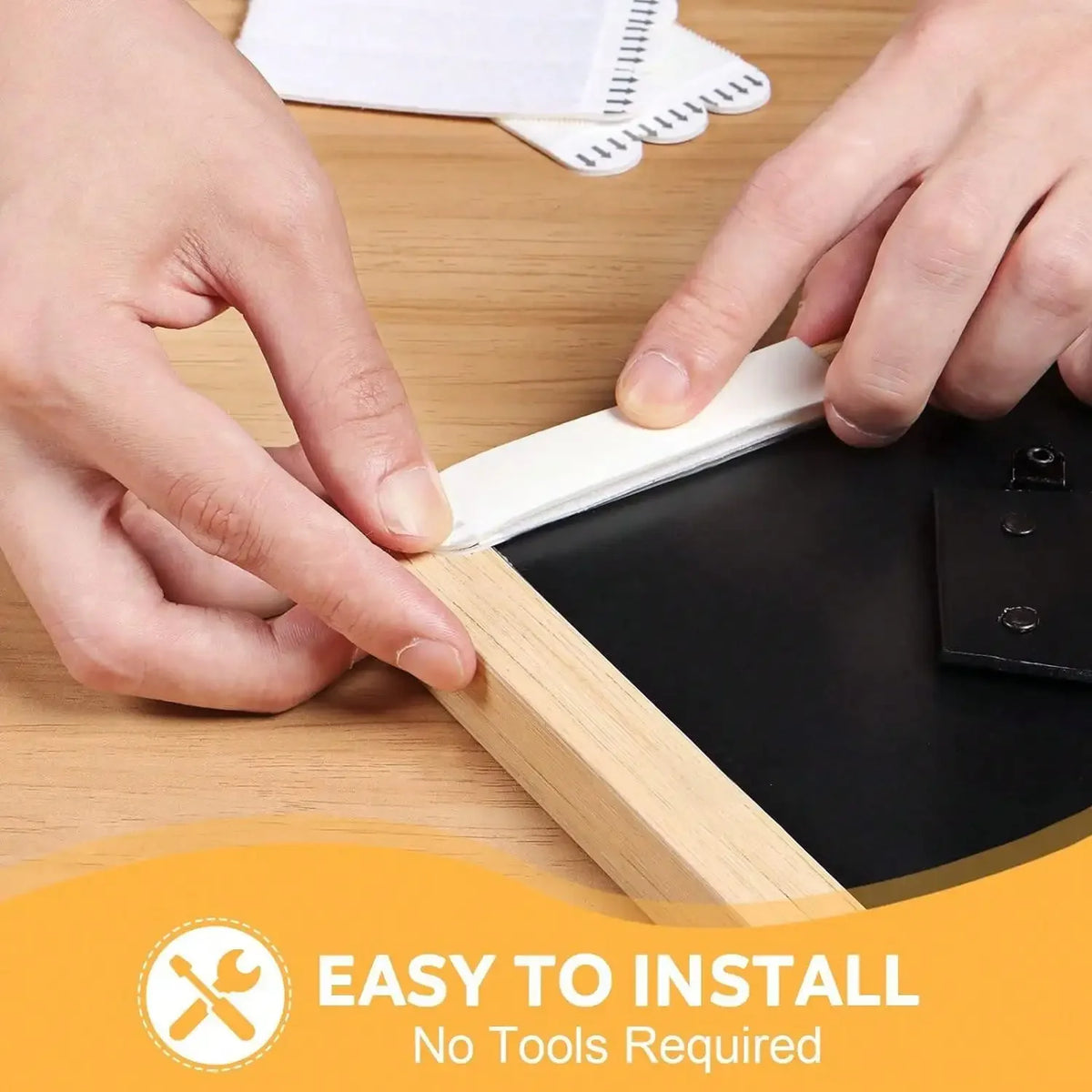 Hands are installing a black frame using white double-sided adhesive strips on a wooden surface.