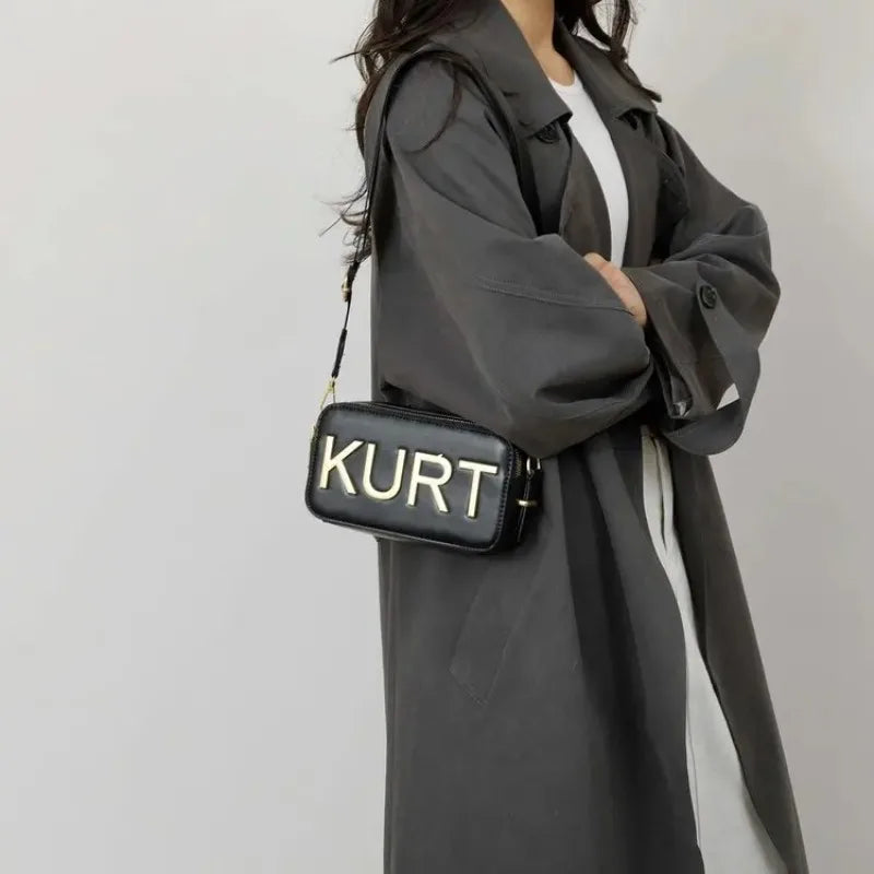 Eine Frau trägt einen grauen Mantel und hält eine elegante schwarze Leder-Totebag mit der Aufschrift KURT.