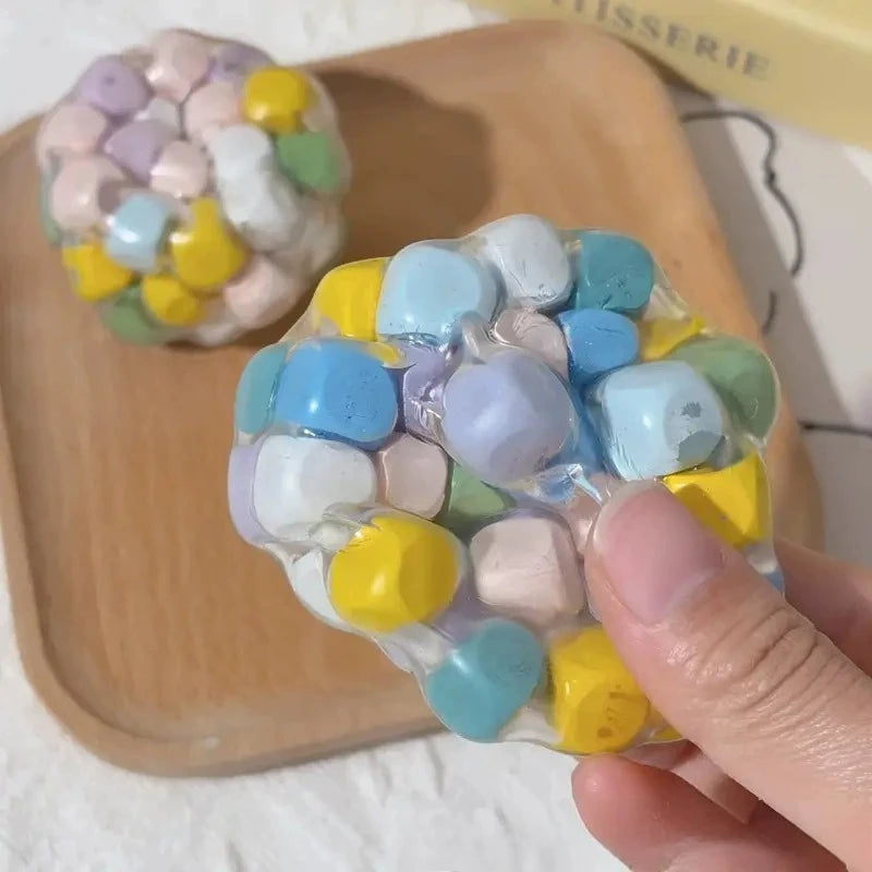 A hand holds a translucent squishy stress ball made of various pastel-colored, soft silicone beads on a wooden tray.