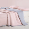 A pink and gray ice silk cooling blanket is draped over a white surface with two light pink pillows nearby.