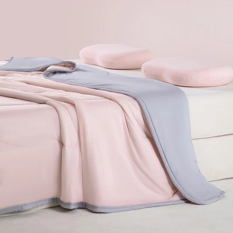A pink and gray ice silk cooling blanket is draped over a white surface with two light pink pillows nearby.